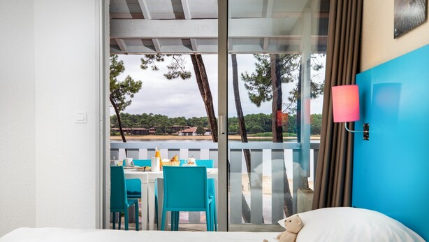 Une chambre d’hôtel avec un lit, une fenêtre donnant sur une plage et des arbres, une table avec des chaises et une lampe.