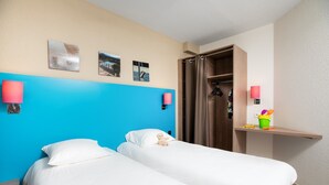Hotelkamer met twee bedden, een blauwe accentmuur, een houten garderobe en een klein plankje met een lamp en een emmer.