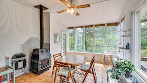 Ein Zimmer mit Holztisch und -stühlen, einem Deckenventilator, einem Kamin und großen Fenstern mit Blick auf Grünflächen.
