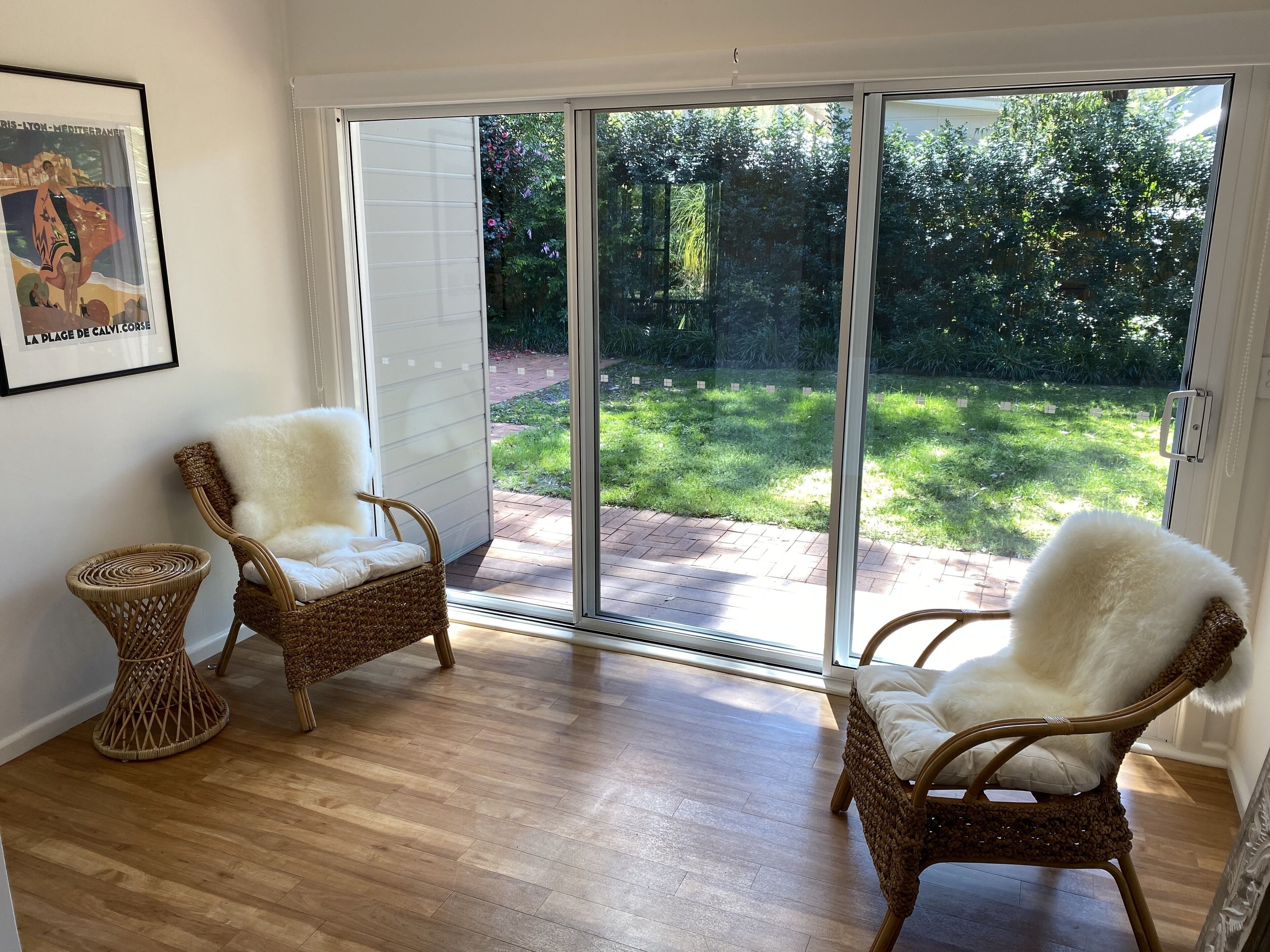 A room with two wicker chairs, a framed picture on the wall, and a sliding glass door leading to a garden.
