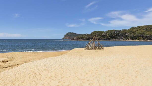 Una platja amb cel blau, mar tranquil·la i una petita construcció de pals.