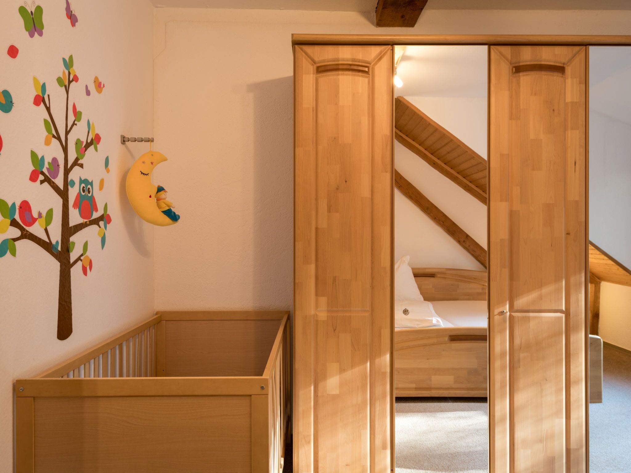 Un berceau en bois orné d’un motif d’arbre peint sur le mur, une armoire en bois et un escalier menant à un lit en mezzanine.
