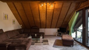 Un loft spacieux avec un plafond haut, des poutres en bois, un canapé d’angle, une petite table basse et une vue dégagée sur l’extérieur grâce à une grande porte-fenêtre coulissante.