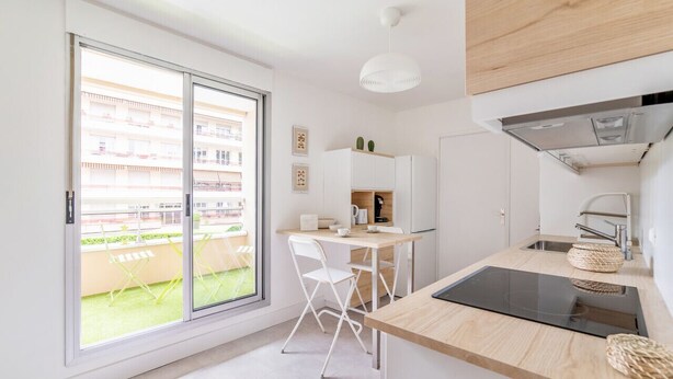 A modern kitchen with a large sliding glass door leading to a balcony, white cabinetry, and a black induction cooktop. - Appartement Mansana - Welkeys (Caluire-et-Cuire)