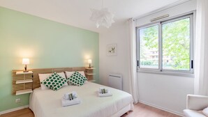 A neatly arranged bedroom with a bed, bedside lamps, a window with a view, and a chair. - Appartement Mansana - Welkeys (Caluire-et-Cuire)