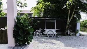Un vélo est garé à côté d’une structure blanche, qui pourrait être une sorte de chariot, dans un espace recouvert de gravier. On aperçoit une clôture noire et de la verdure en arrière-plan.