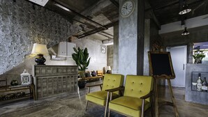 Une pièce avec un mur en pierre, un meuble en bois, un fauteuil vert, un chevalet en bois et une horloge au mur.