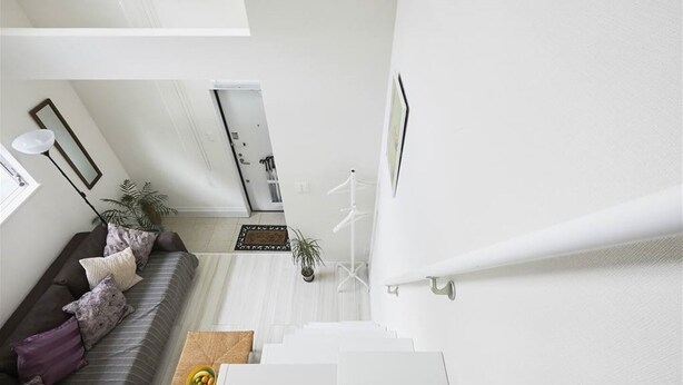 A modern interior with a staircase leading to an upper level, a sofa with patterned cushions, a small table with a bowl of fruit, and a potted plant.