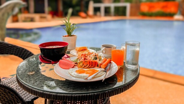 Una mesa de comedor con comida, un vaso de jugo de naranja y un vaso de agua junto a una piscina.