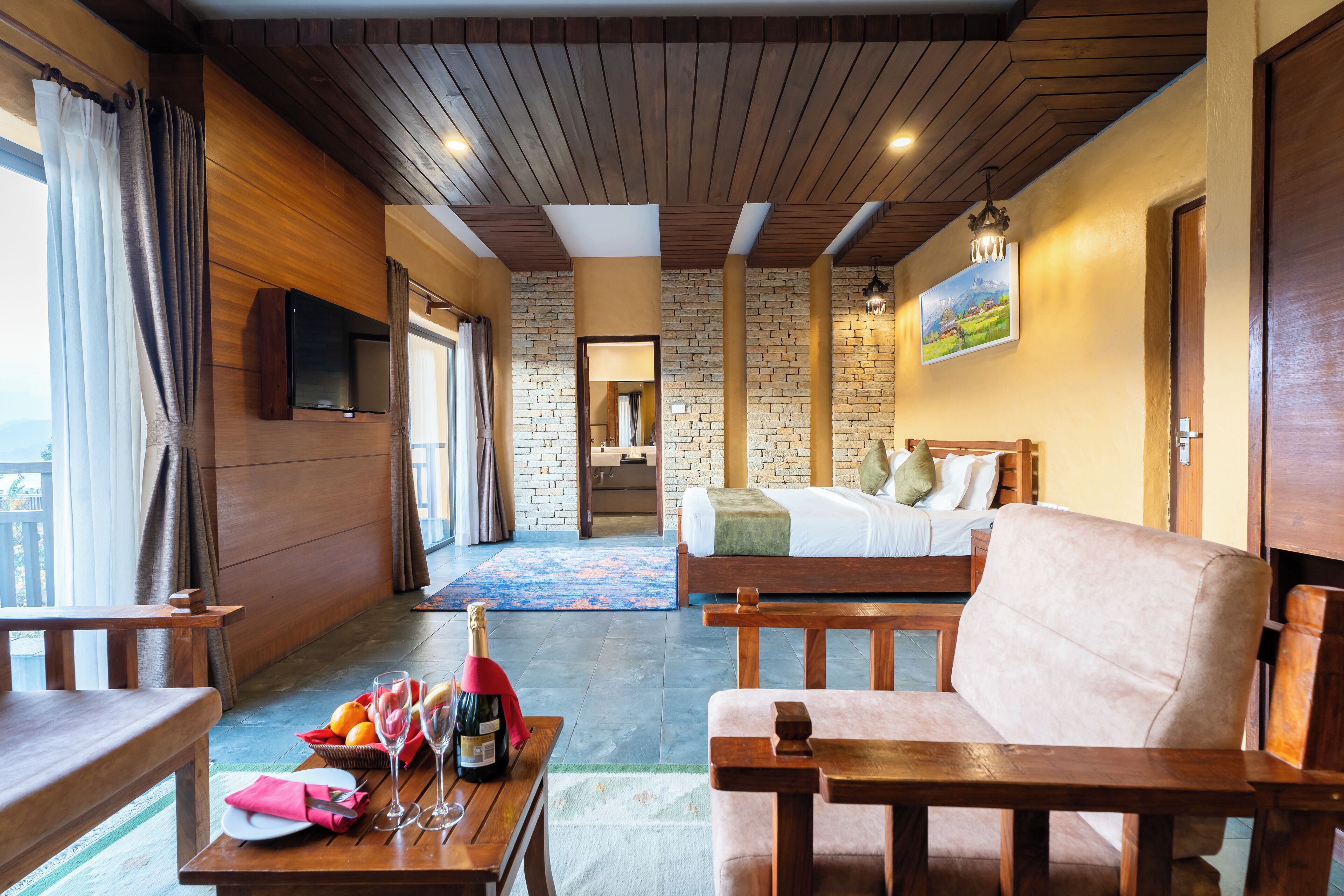 A hotel room with a wooden ceiling, brick walls, a bed, a sofa, a table with a bottle of champagne, and a TV mounted on the wall.