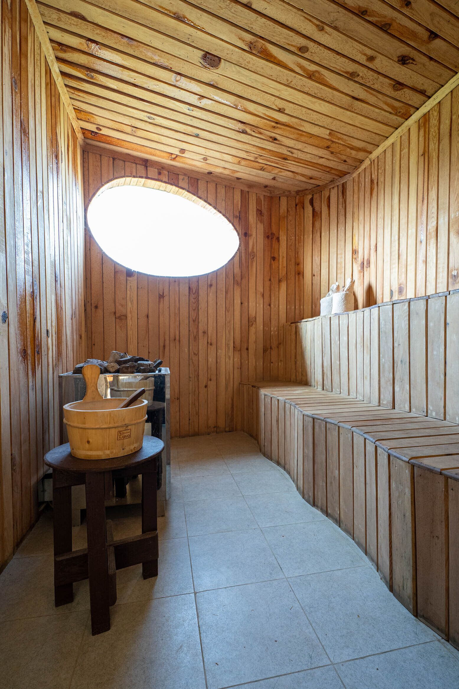 A wooden cabin interior with a stone fireplace, a wooden table with a bucket, and benches along the walls.