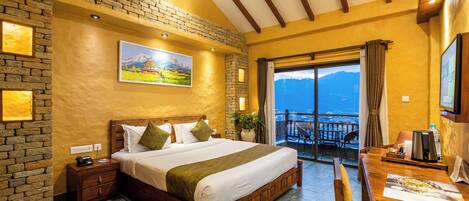 A bedroom with a large bed, a wooden desk, a balcony with a view, and a stone wall.