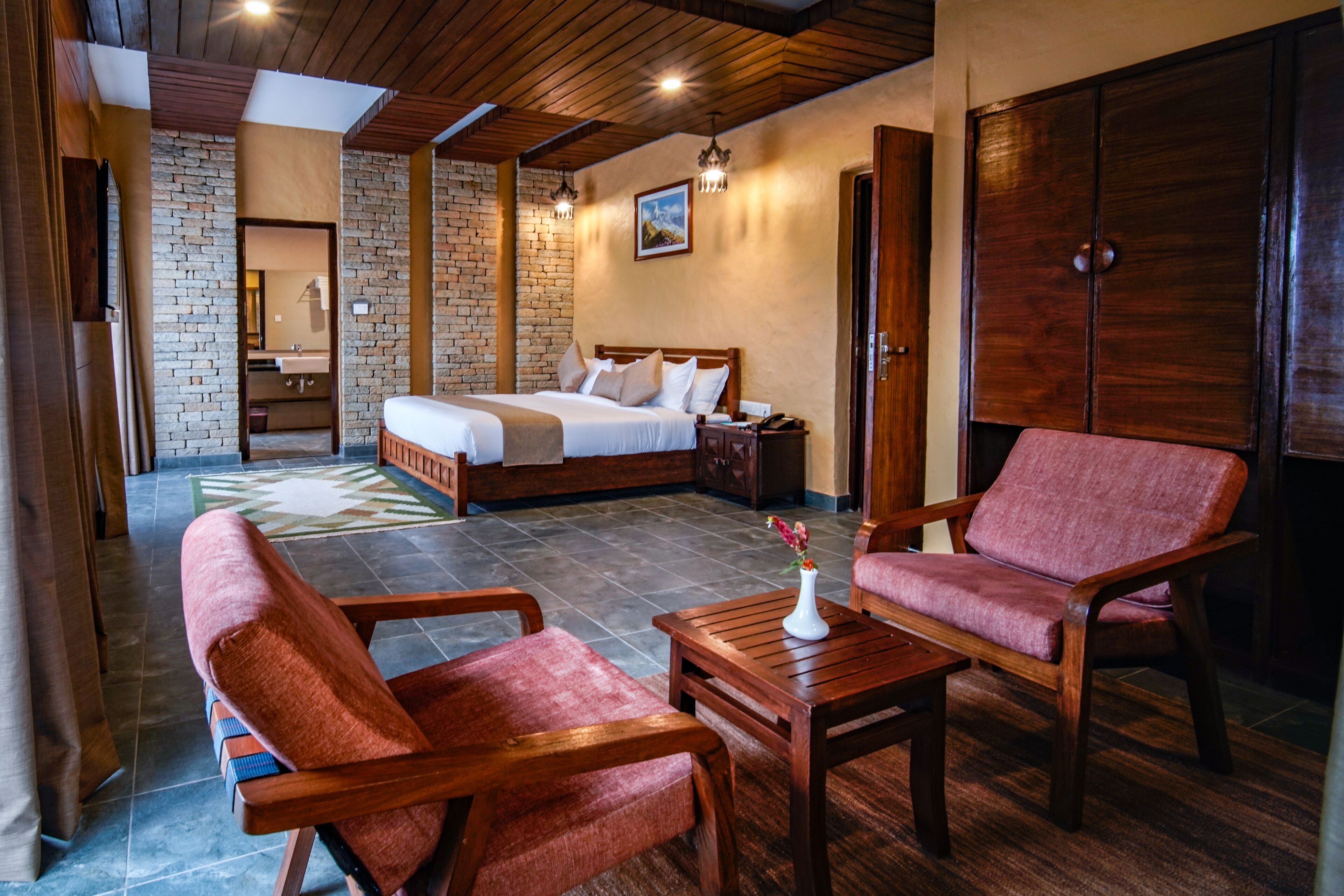 A hotel room with a bed, a wooden wardrobe, a seating area with a table and chairs, and a bathroom visible in the background.