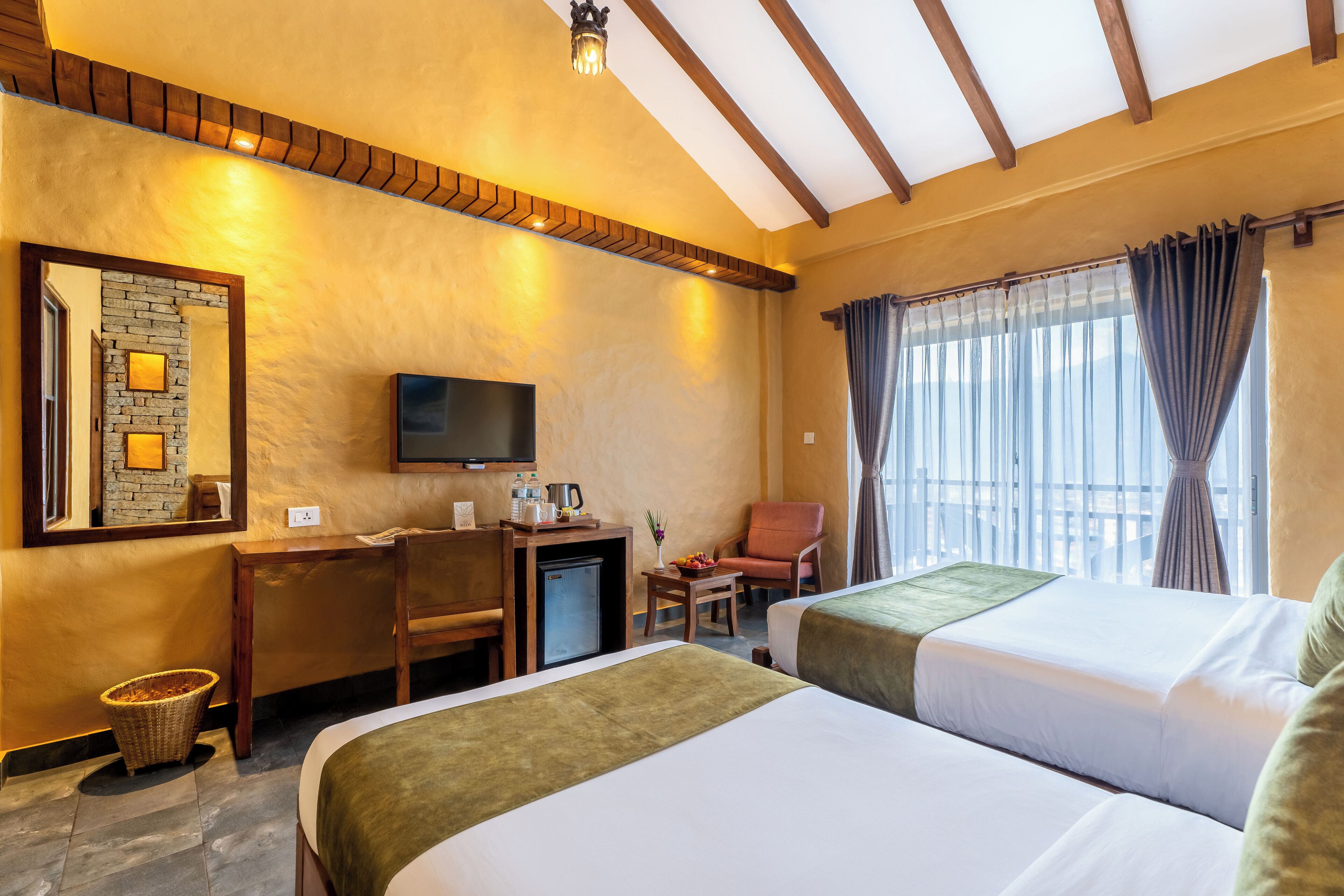 Een hotelkamer met twee bedden, een bureau met televisie, een klein tafeltje met een schaal fruit, een stoel en een raam met gordijnen.