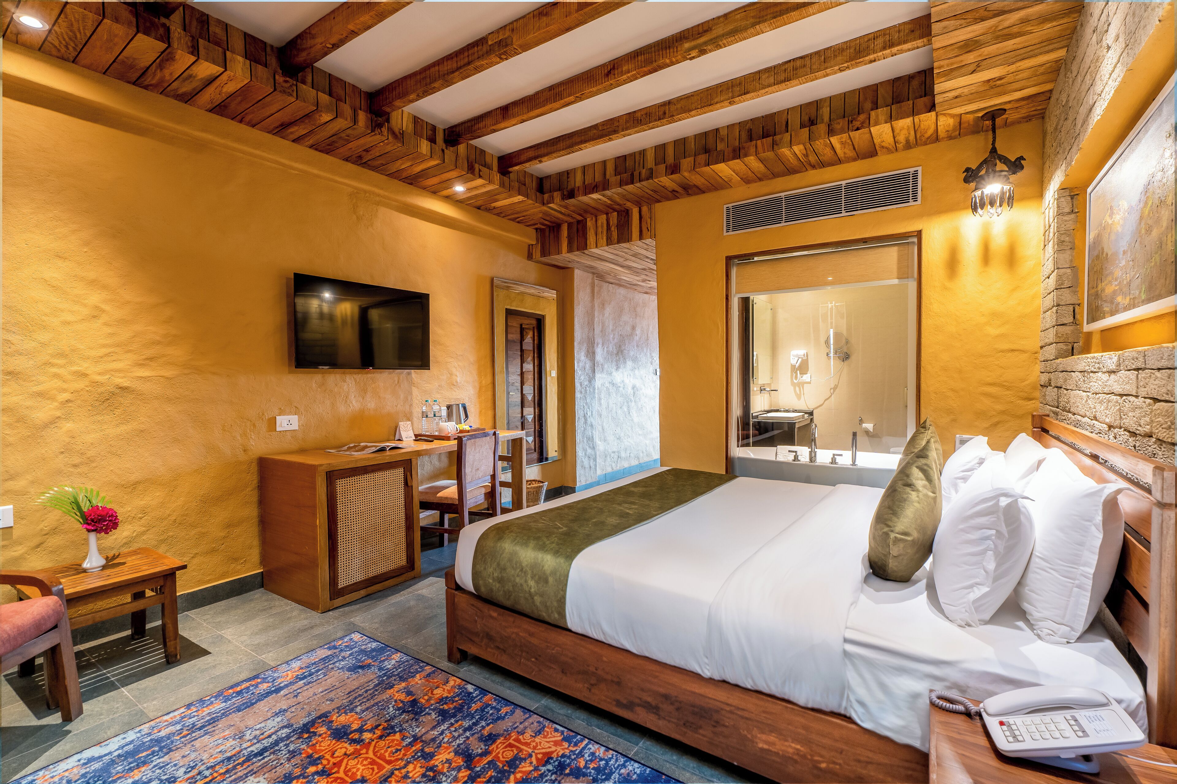 A hotel room with a wooden ceiling, a bed with white bedding, a desk with a chair, a television, and a bathroom with a large mirror.
