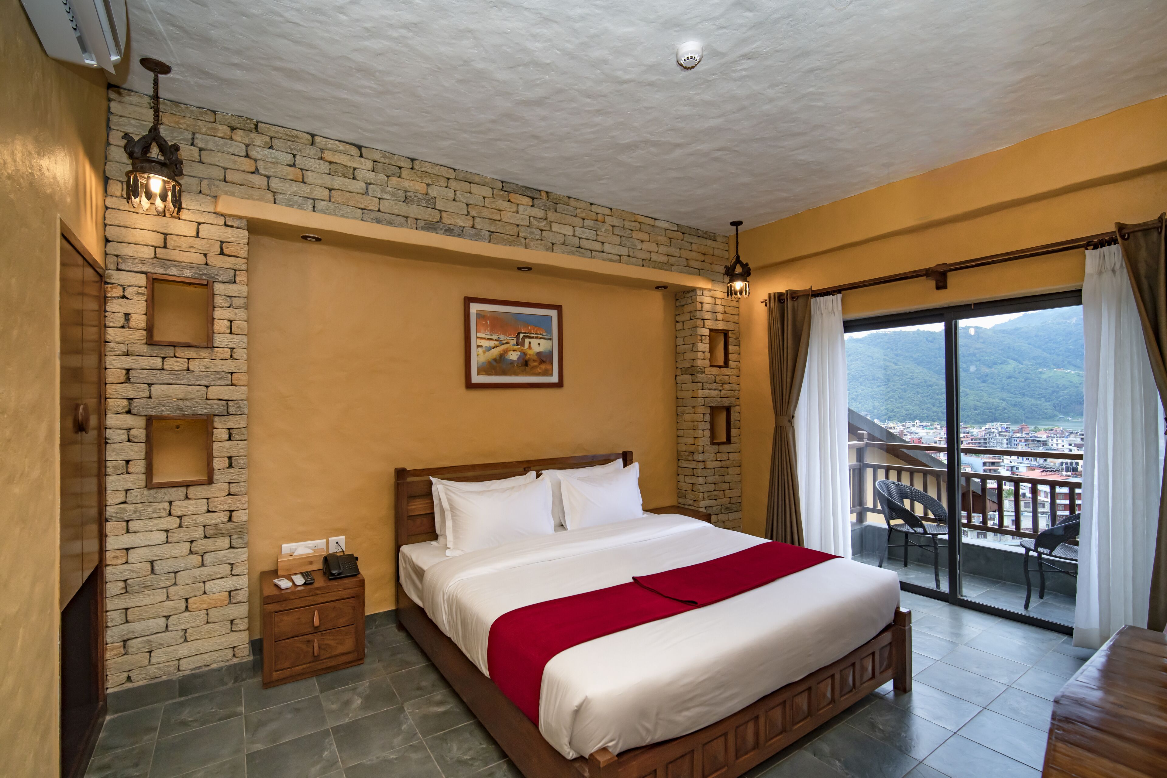 Habitación de hotel con una cama grande, una mesita de noche de madera, una pequeña mesa, un muro decorativo de ladrillo, un cuadro y un balcón con vistas.