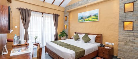 A hotel room with a wooden bed, a desk, a chair, a large window with curtains, a painting on the wall, and a stone accent wall.