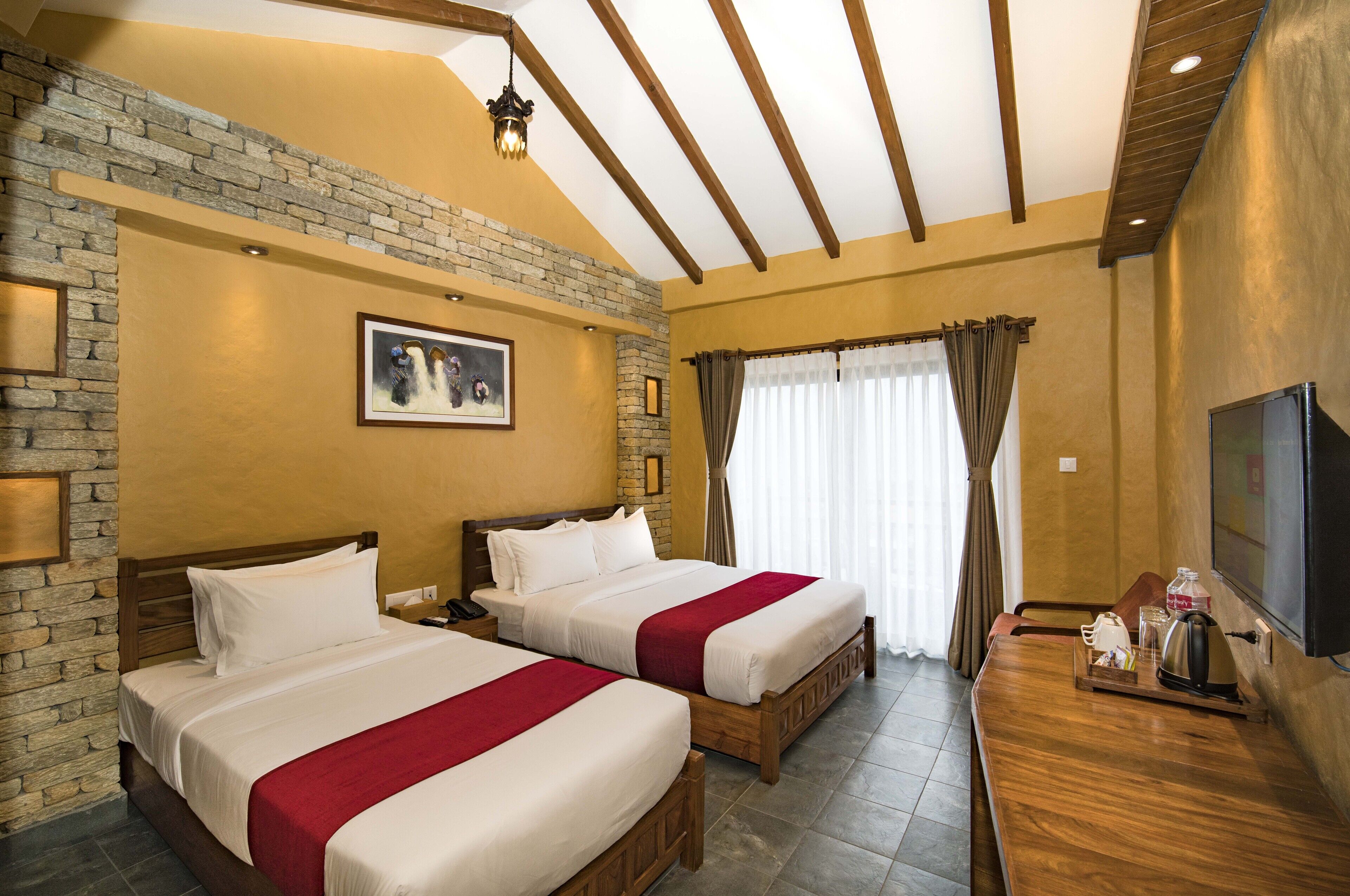 A hotel room with two beds, a stone accent wall, a mounted TV, and a wooden desk with a tray of items.