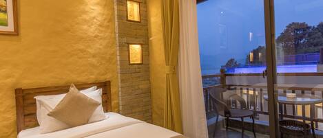 Habitación de hotel con cama, pared de piedra, balcón con sillas y vista al mar.