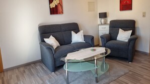 A modern living room with a grey sofa set, a glass coffee table, and two armchairs. There are two wall-mounted artworks, a lamp on a side table, and a small cabinet. - Ferienwohnung 3 - Ferienwohnung Haus Bikker (Ihlow)