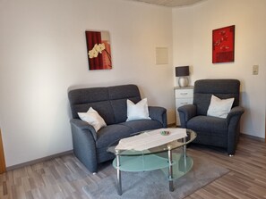 A modern living room with a grey sofa set, a glass coffee table, and two armchairs. There are two wall-mounted artworks, a lamp on a side table, and a small cabinet. - Ferienwohnung 3 - Ferienwohnung Haus Bikker (Ihlow)