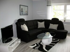 A modern living room with a black sectional sofa, a glass coffee table, a flat-screen TV on a white stand, a framed artwork on the wall, and a window with a view of greenery. - Ferienwohnung 3 - Ferienwohnung Haus Bikker (Ihlow)