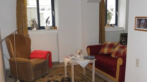 A living room with a brown armchair, a red sofa, a small white table, and a floor lamp. There are windows with curtains, a radiator, and a framed picture on the wall. - Sleep well apartment, Germany (Görlitz)