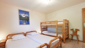 Une chambre avec deux lits simples et un lit superposé, des meubles en bois, une petite table et un tableau au mur.