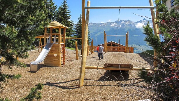 Un terrain de jeux avec un toboggan et des balançoires, entouré d’arbres et offrant une vue sur la montagne.