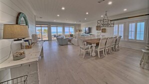 A spacious living area with a dining table, white furniture, and large windows offering a view of the beach.