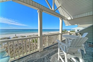 Un espacio al aire libre techado con vista a la playa, sillas blancas y una mesa.