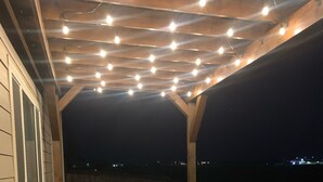 Une terrasse extérieure avec des meubles en osier et une pergola illuminée de guirlandes lumineuses.