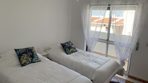 A bedroom with two beds, white bedding, a white ceiling fan, and a window with a view of buildings outside. - Pinhal Do Golf: Superb 2-bedroom apartment, renovated in 2020 near Marina (Quarteira)