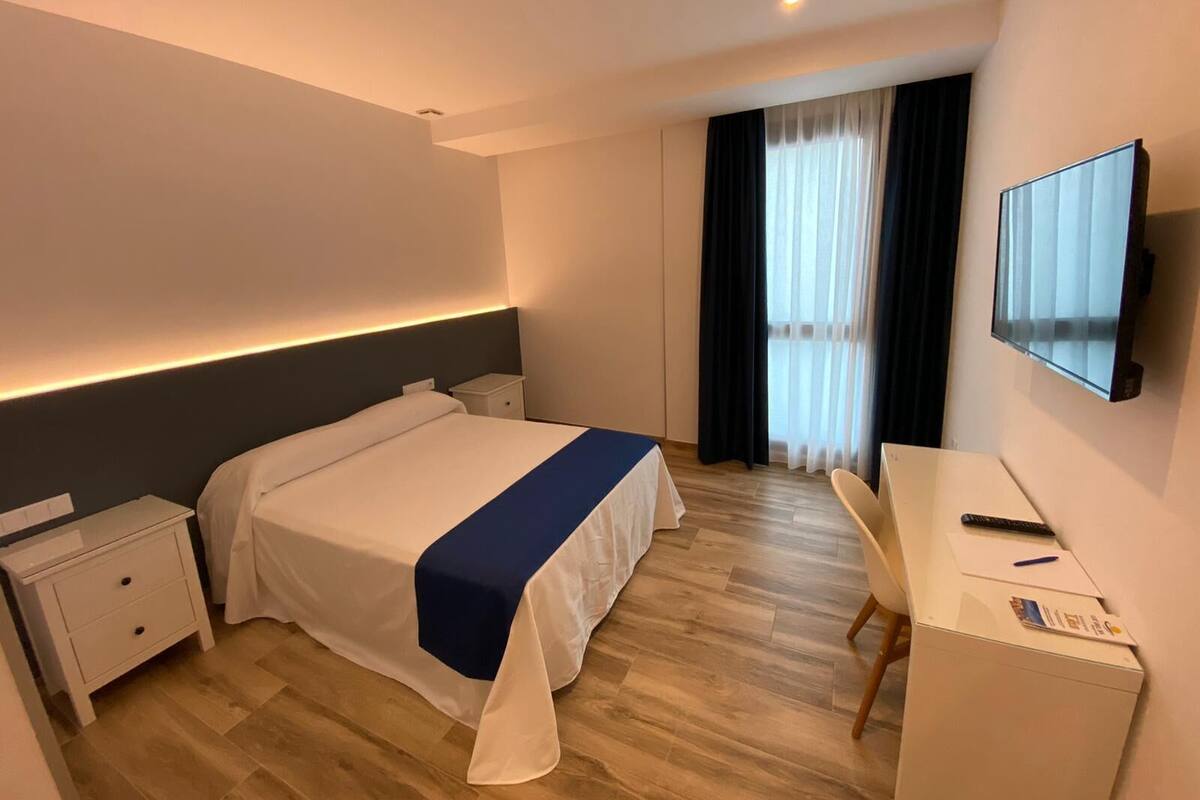 A hotel room with a bed, bedside tables, a desk, a chair, a TV, and a window with curtains.