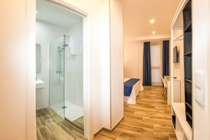A hotel room with a glass shower, a bed with a blue bedspread, a desk with a chair, and a TV on the wall. - Hostal Luz del Sol Barbate (Barbate)
