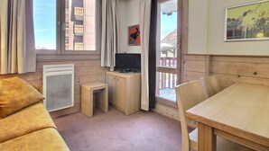 Una habitación con piso de madera, un sofá, una mesa, un televisor y una ventana con cortinas.
