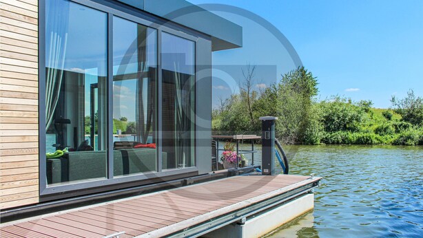 Ein modernes Hausboot mit großen Fenstern, einem Holldeck und Blick auf einen Fluss und Grünflächen.