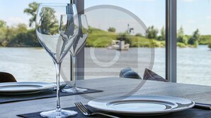 Ein Tisch mit zwei Weingläsern, Tellern und Besteck, mit Blick auf ein Gewässer, in dessen Hintergrund sich Grünflächen und ein Steg befinden.
