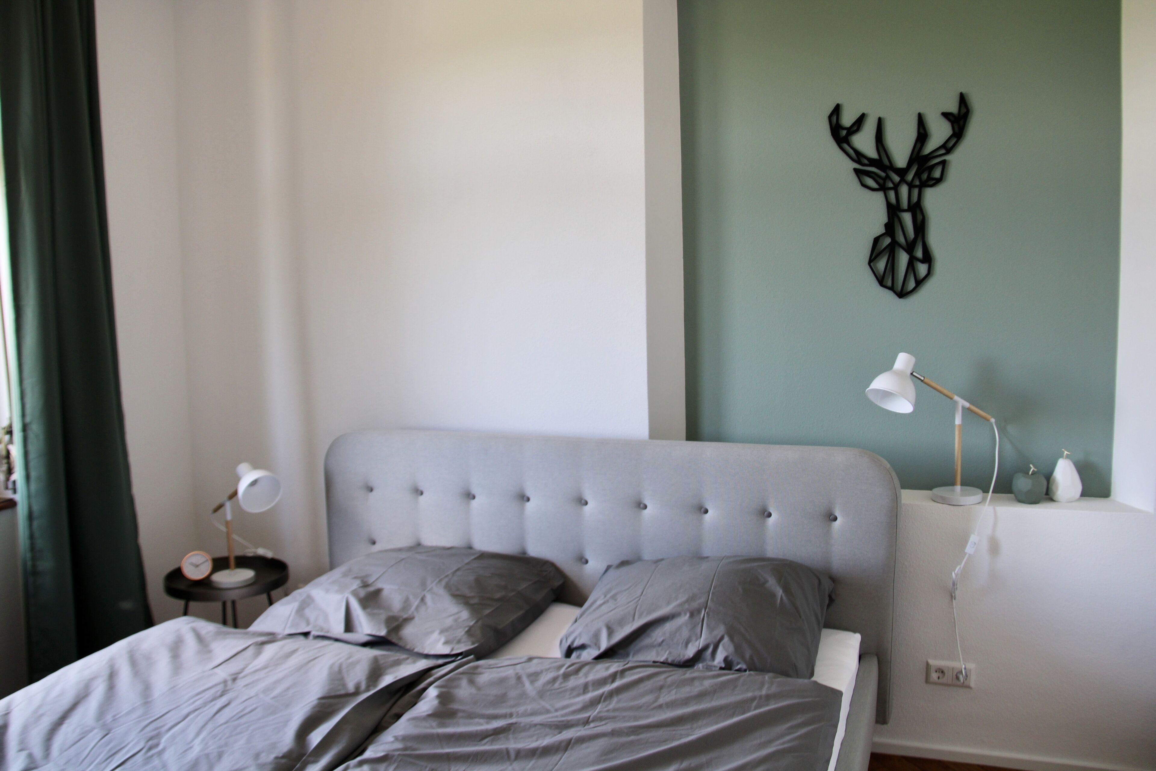 A bedroom with a tufted headboard, grey bedding, a wall-mounted deer head decoration, a bedside lamp, and a small table with a clock.