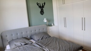 Un lit bien fait, avec une literie grise, une tête de cerf décorative accrochée au mur et une armoire blanche aux poignées argentées.