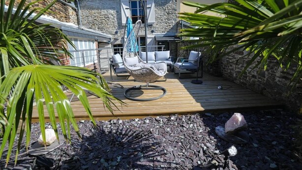Una terraza de madera con mobiliario exterior, incluyendo una hamaca colgante y sillones reclinables, frente a un edificio de piedra con techo inclinado.