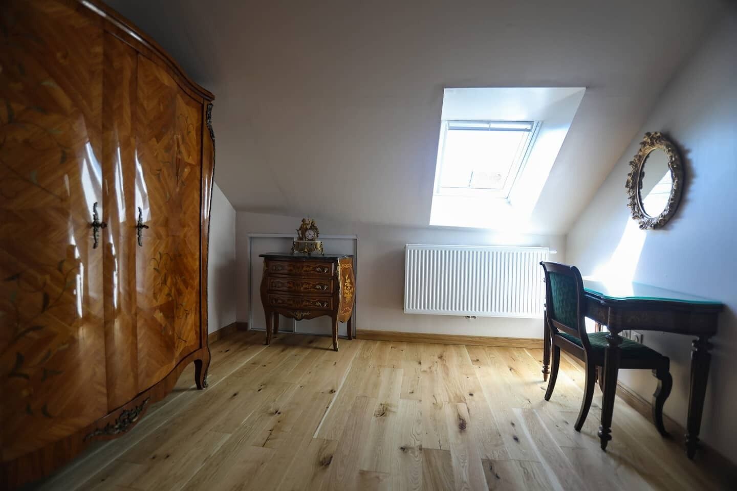 A room with a wooden floor, a dresser, a desk, a chair, a radiator, and a skylight.
