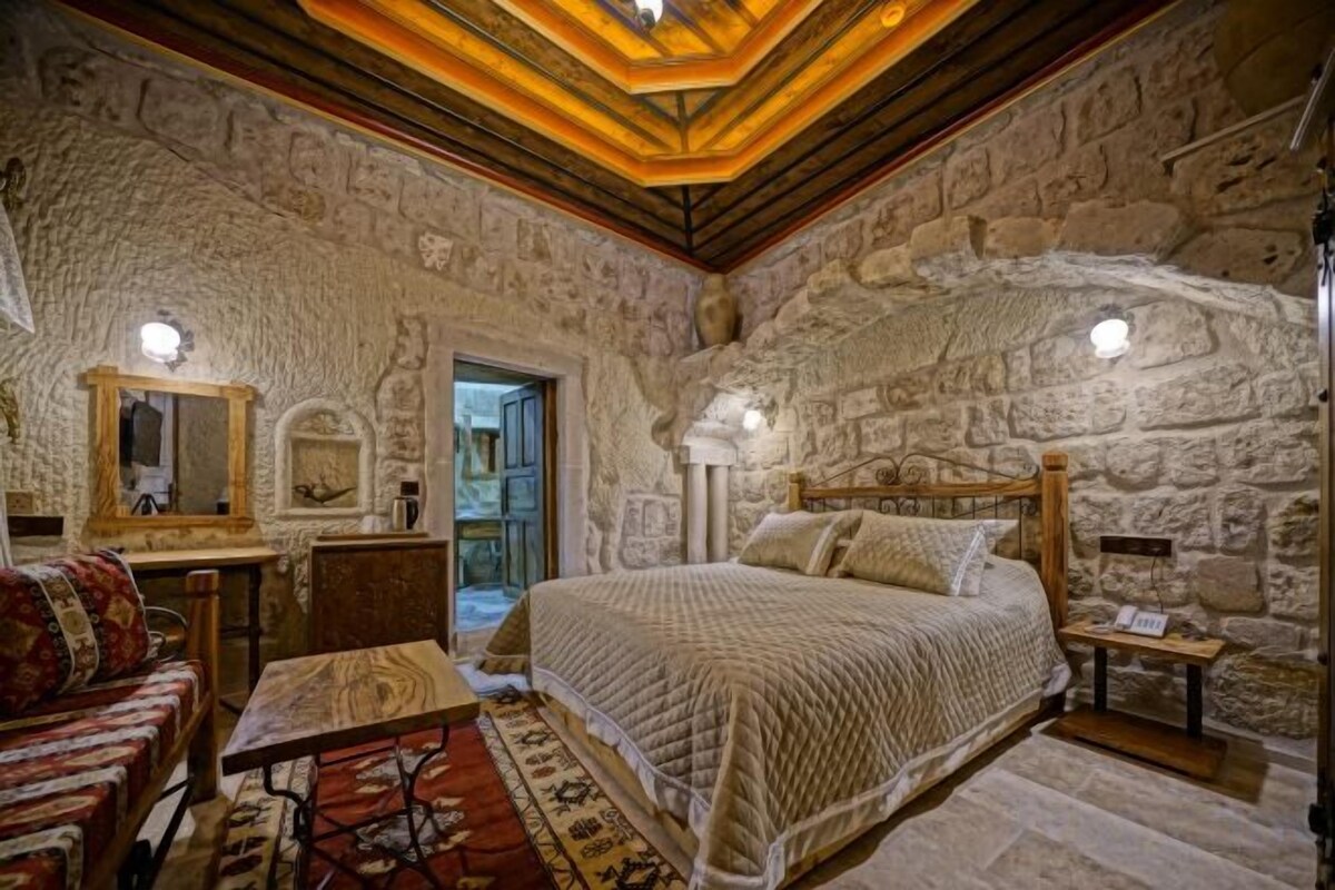 A stone-walled bedroom with a bed, a nightstand, a lamp, and a mirror.