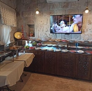 A buffet setup with various dishes, a television mounted on the wall, and a stone wall background. - Cappadocia Cave Lodge (Nevsehir)