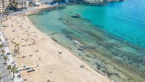 An aerial view of a beach with clear turquoise waters, a sandy shore, and buildings in the background.