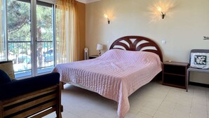 A bedroom with a bed, a chair, a nightstand, a lamp, and a window with curtains. - Charming 2-Room Apartment Near Sea in Argelès-sur-Mer (Argelès-sur-Mer)