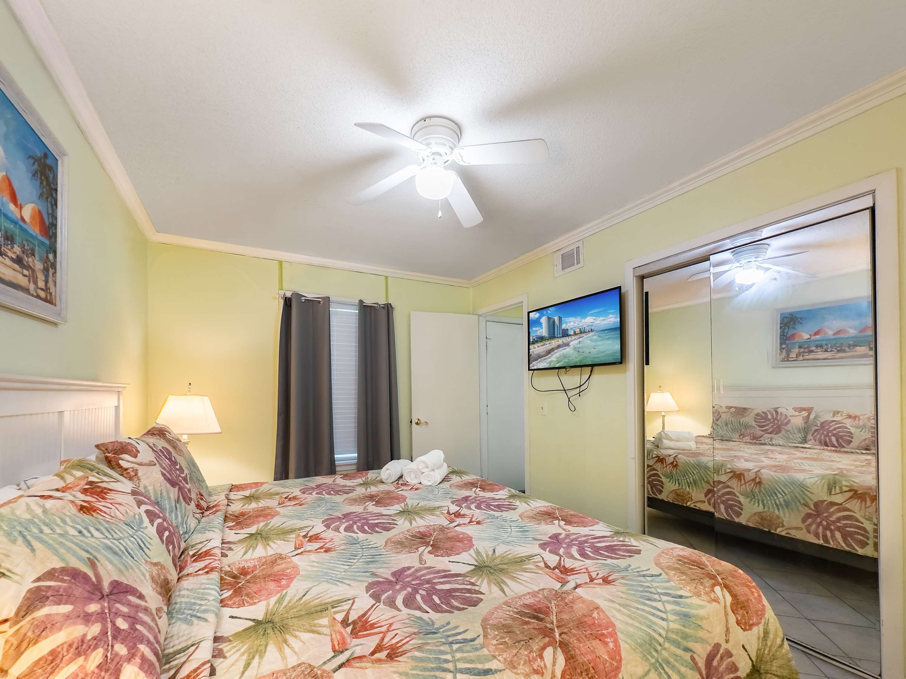 Habitación de hotel con cama, ventilador de techo, televisor y puerta que da a un baño.
