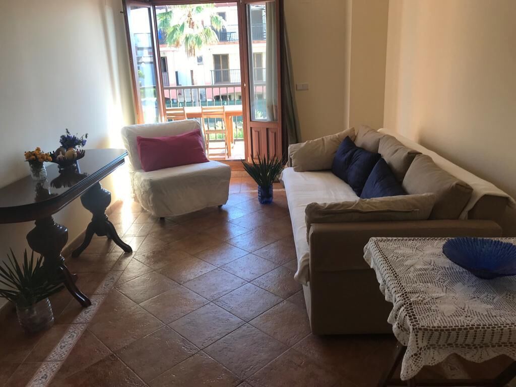 Ein Wohnzimmer mit einem weißen Sofa, einem beigen Sofa, einem kleinen Tisch mit Blumen und Blick auf einen Balkon mit Geländer.