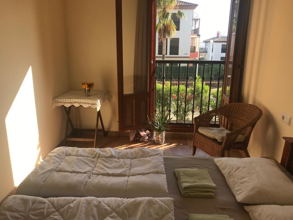 Een slaapkamer met twee bedden, een tafeltje met een bloevas, een rieten stoel en uitzicht op een balkon met een hekwerk en begroening.