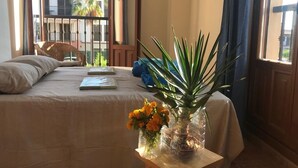 Une chambre à coucher avec un lit, une petite table ornée d’une plante et de fleurs, et une vue sur des immeubles par la fenêtre.